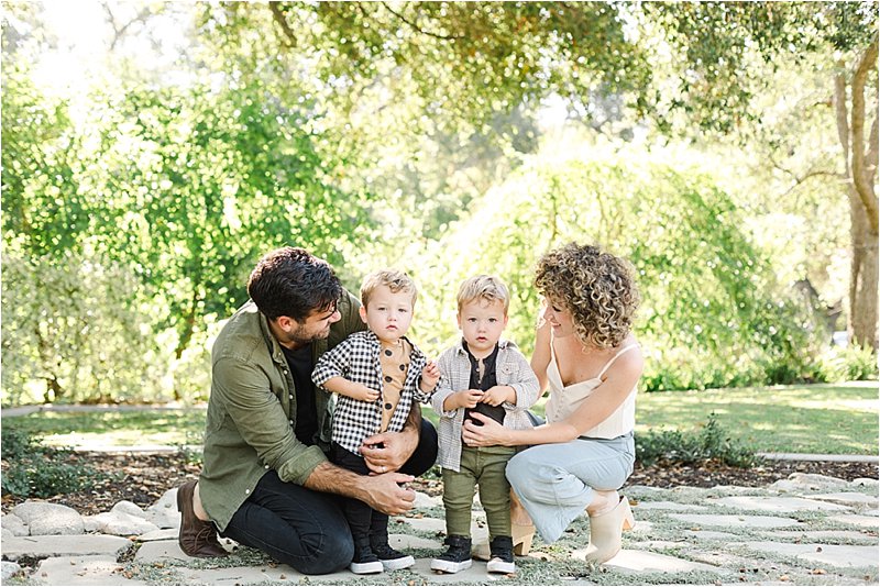 Parents squat down to pose with their 1 year old twin sons under a canopy of trees during their fall family photos.