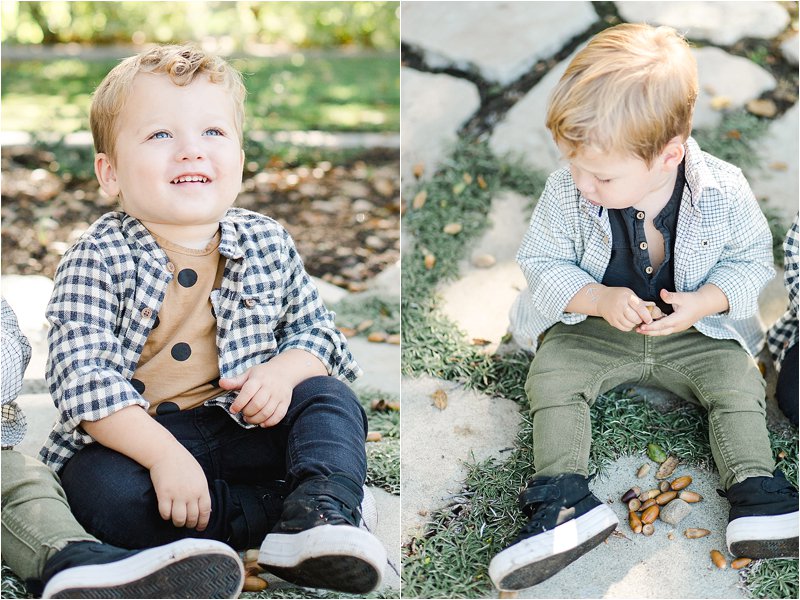 Image on the left shows one twin looking up to the sky + smiling, wearing a brown t-shirt with brown polka dots with a black and white checkered button down layered on top.  Paired with black jeans and black high top sneakers.  Image on the right shows the other twin sitting on the flagstone looking for acorns, wearing a black t-shirt with a cream and taupe flannel shirt layered on top.  Paired with olive green pants and black high top sneakers, showing inspiration for fall family photo outfits.
