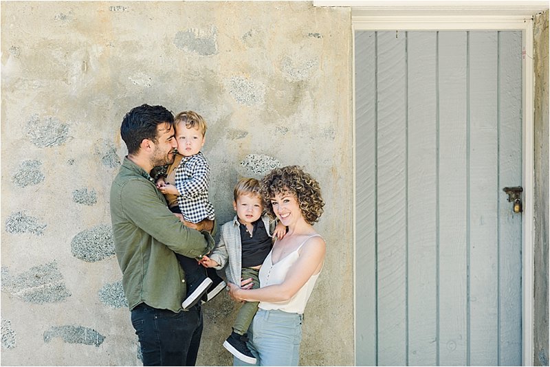 Interactive family portrait as parents hold their 2 year old twin sons in front of grey-blue door.