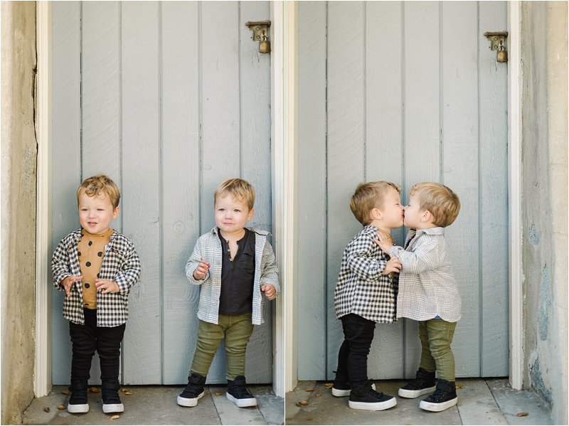 Two year old identical twin boys kiss each other while standing in front of a grey-blue door during their family photos in Pasadena, CA.