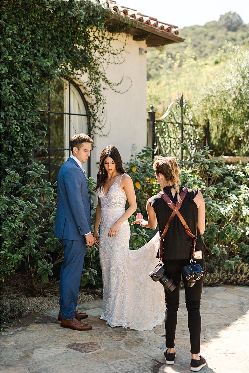 Wedding photographer Tiffany J Photography, wearing a black dress top and pants with a leather harness holding 2 cameras, directs the bride and groom during their wedding portraits.