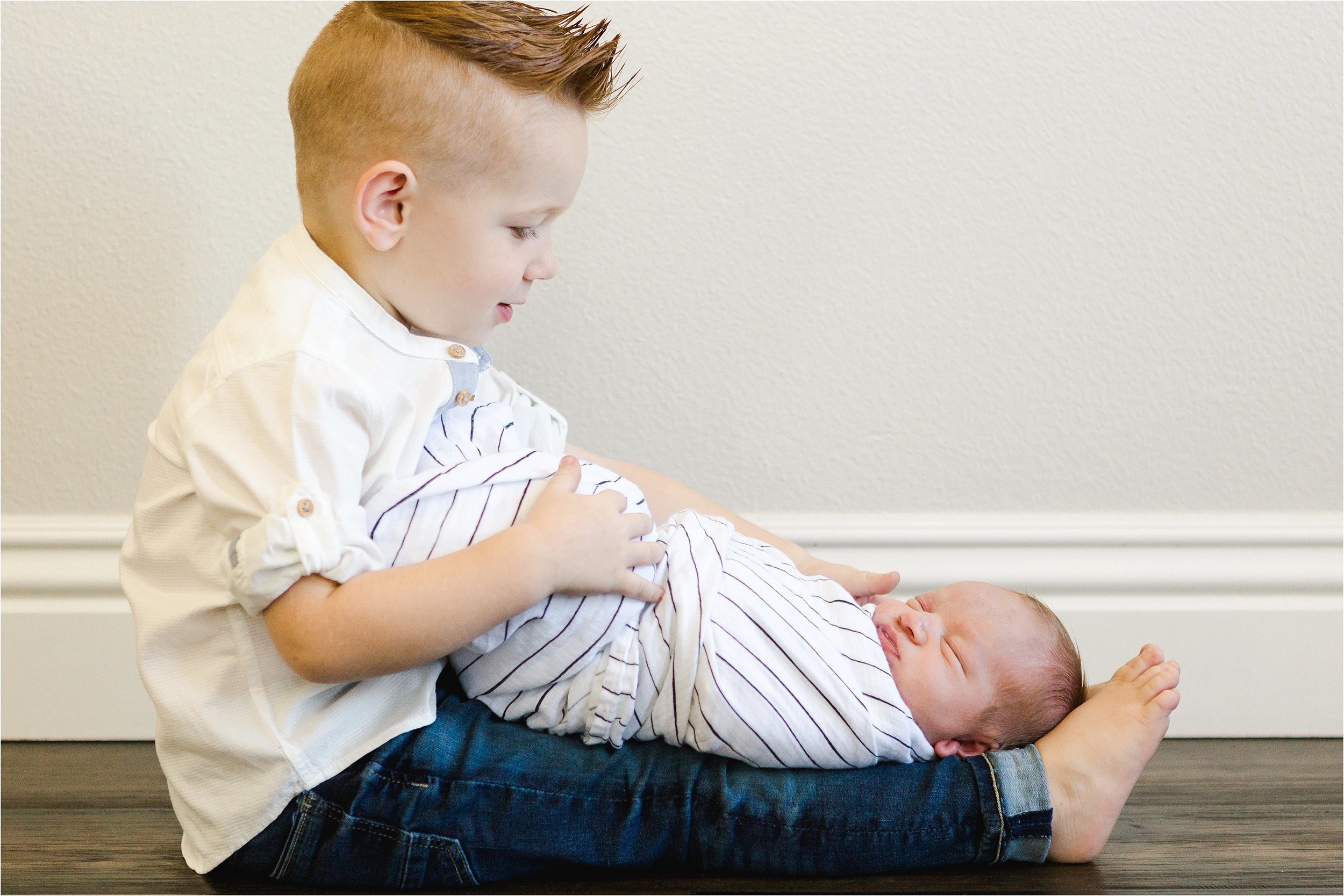 Red-readed big brother sits on the ground with his baby brother lying on his lap.