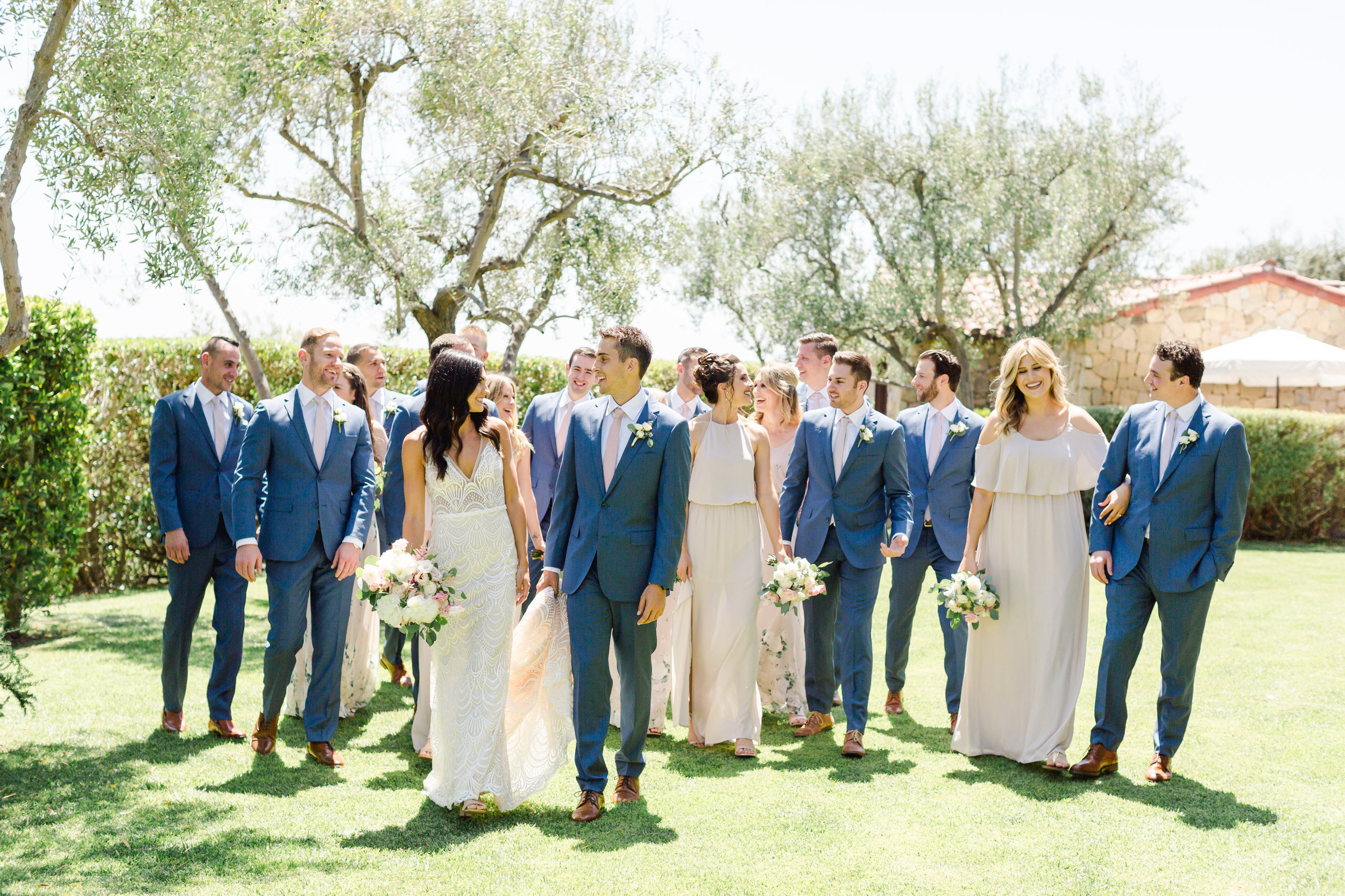 Bride and groom walking with their bridal party on a lawn surrounded by olive trees.