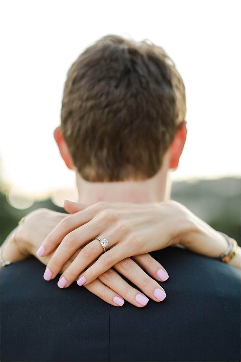 Engagement ring detail shot with woman's hands draped over her fiance's neck showcasing her solitaire engagement ring.