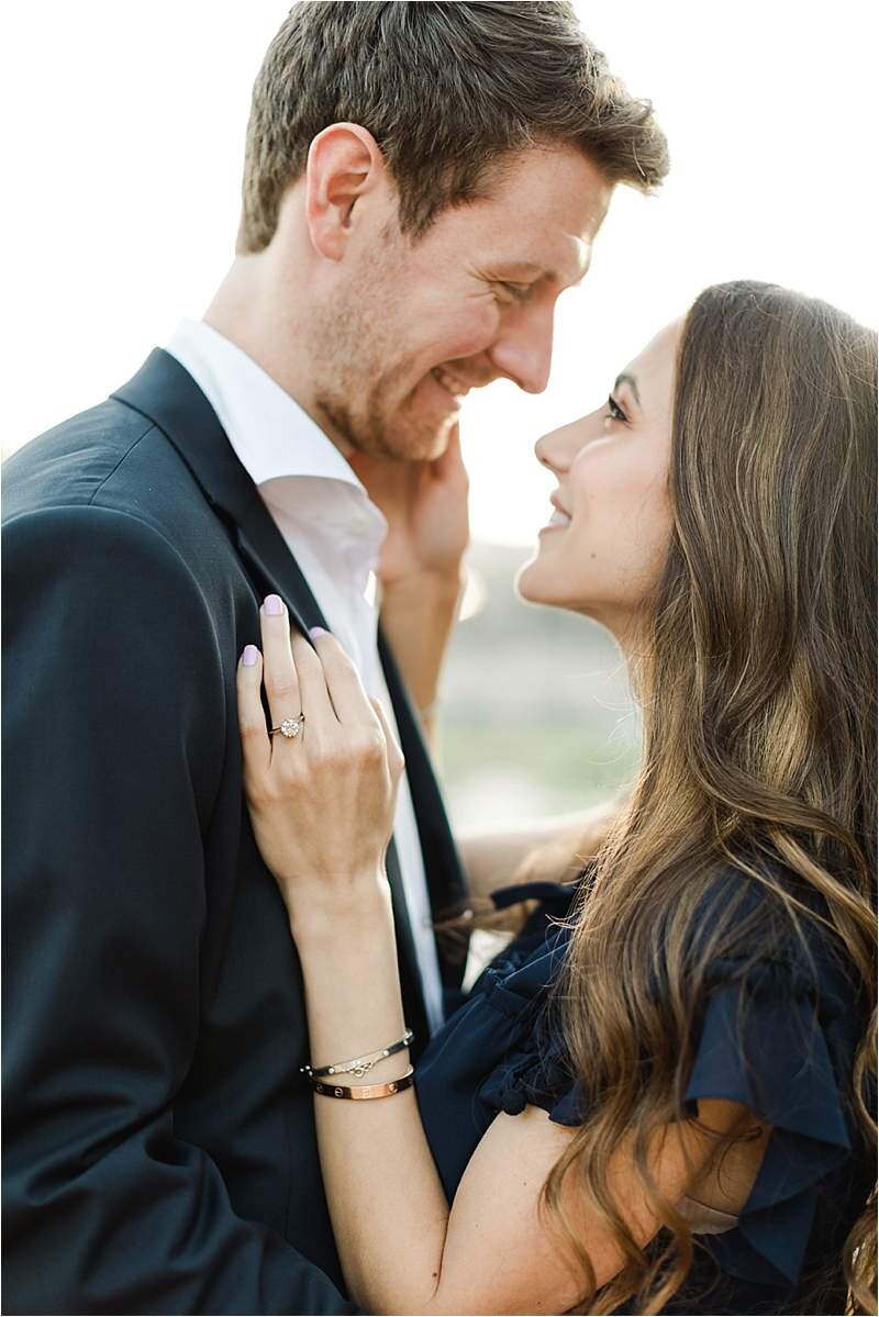 Fiance's smile at each other as she delicately places her hand on his lapel, showcasing her engagement ring following their surprise proposal near Royce Hall at UCLA.