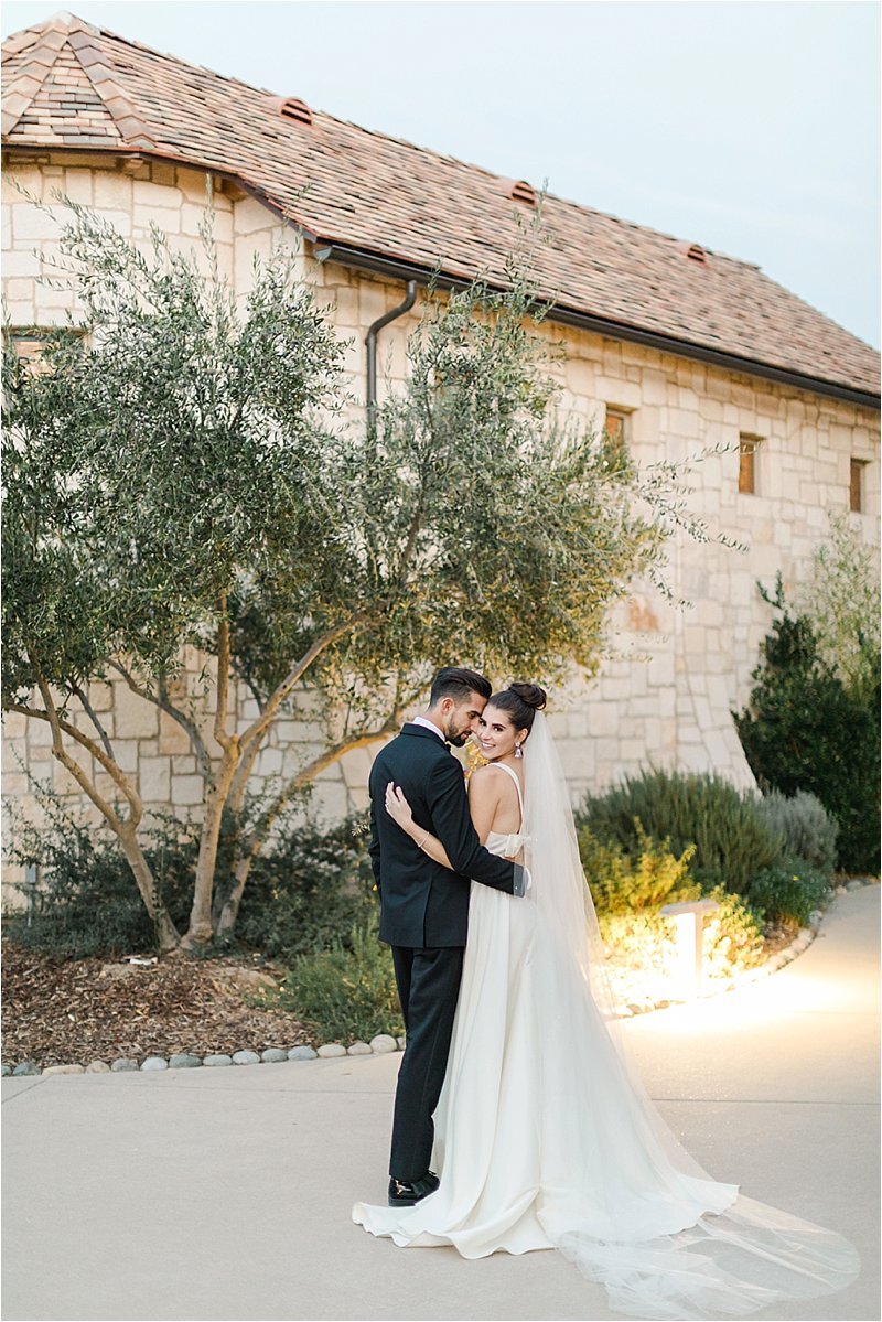 The bride and groom embrace in front of a stone chapel surrounded by olive trees as she smiles over her shoulder and he rests his head on her temple. 