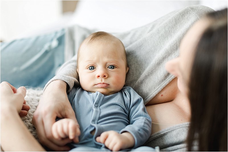Blue eyed baby boy stares straight at the camera while being propped up against his Mother.