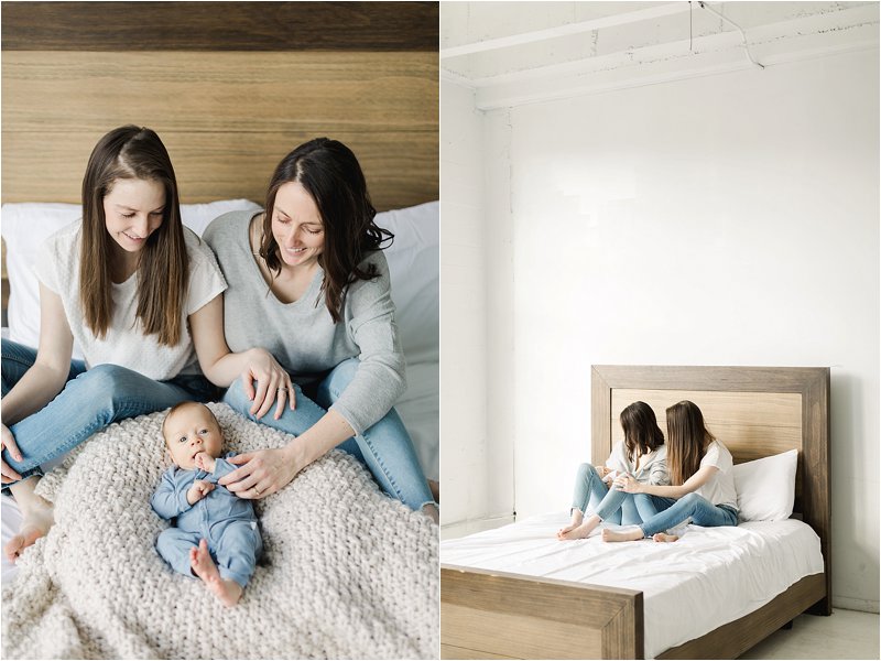 Lesbian couple sit next to each other on a bed while smiling at their newborn son.