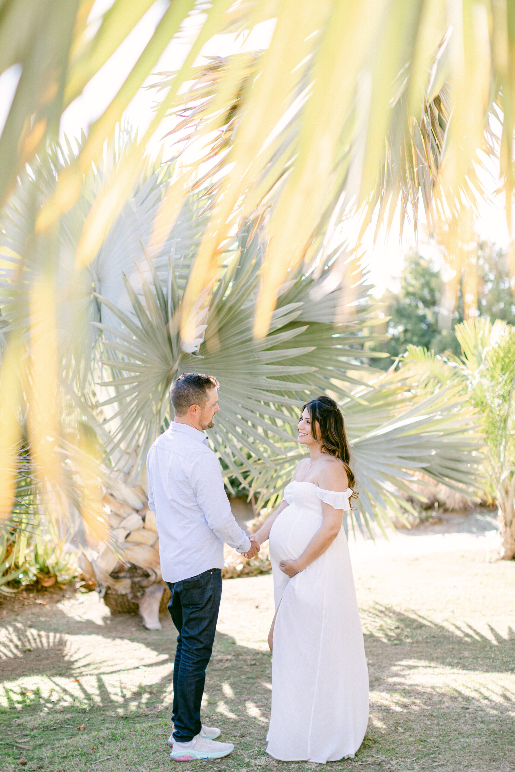 Husband and wife hold hands and smile at each other under palm trees during their maternity photography session at the LA Arboretum.