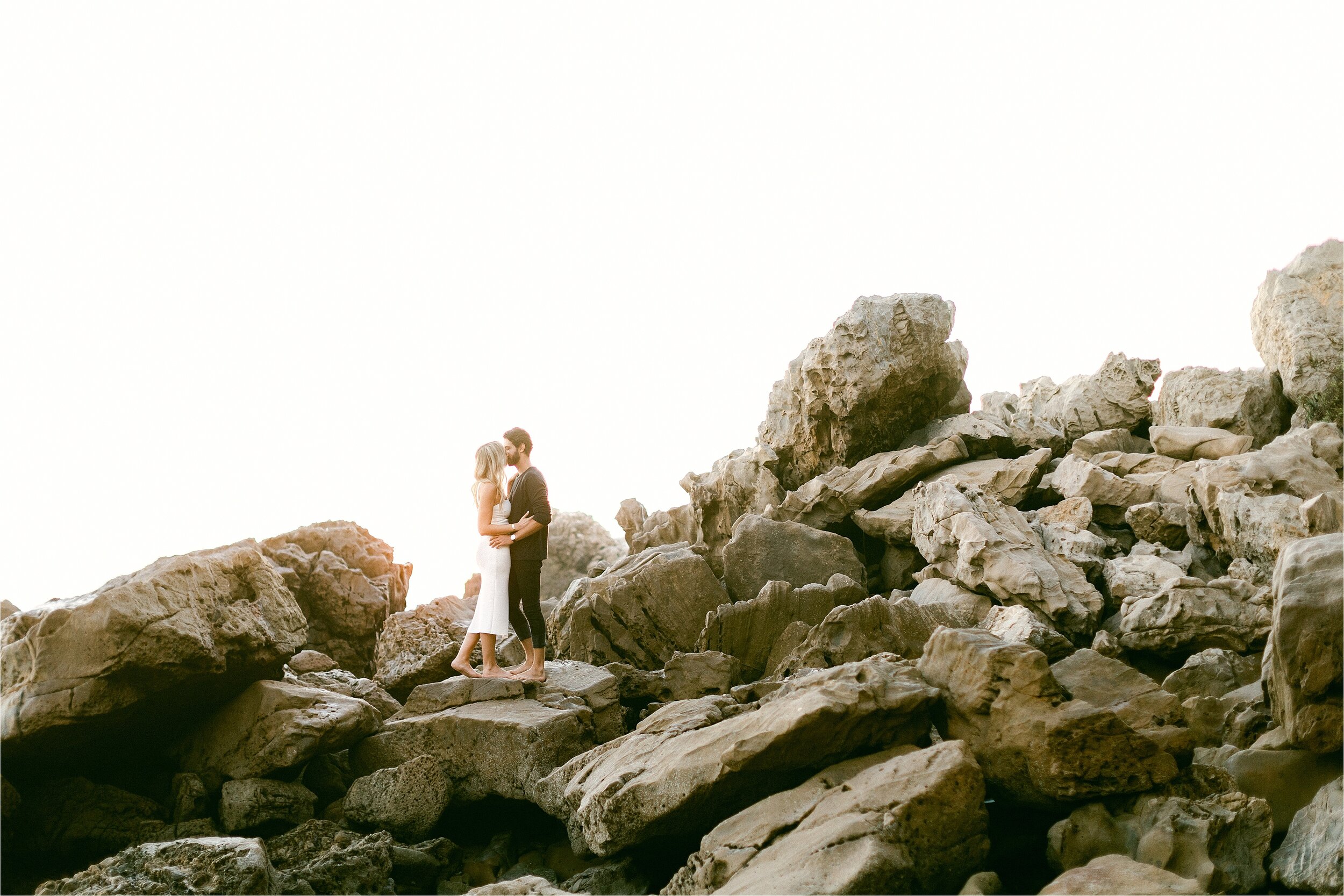 blonde female in white dress kisses her fiance in black jeans and dark tshirt while standing on large rock embankment at the beach