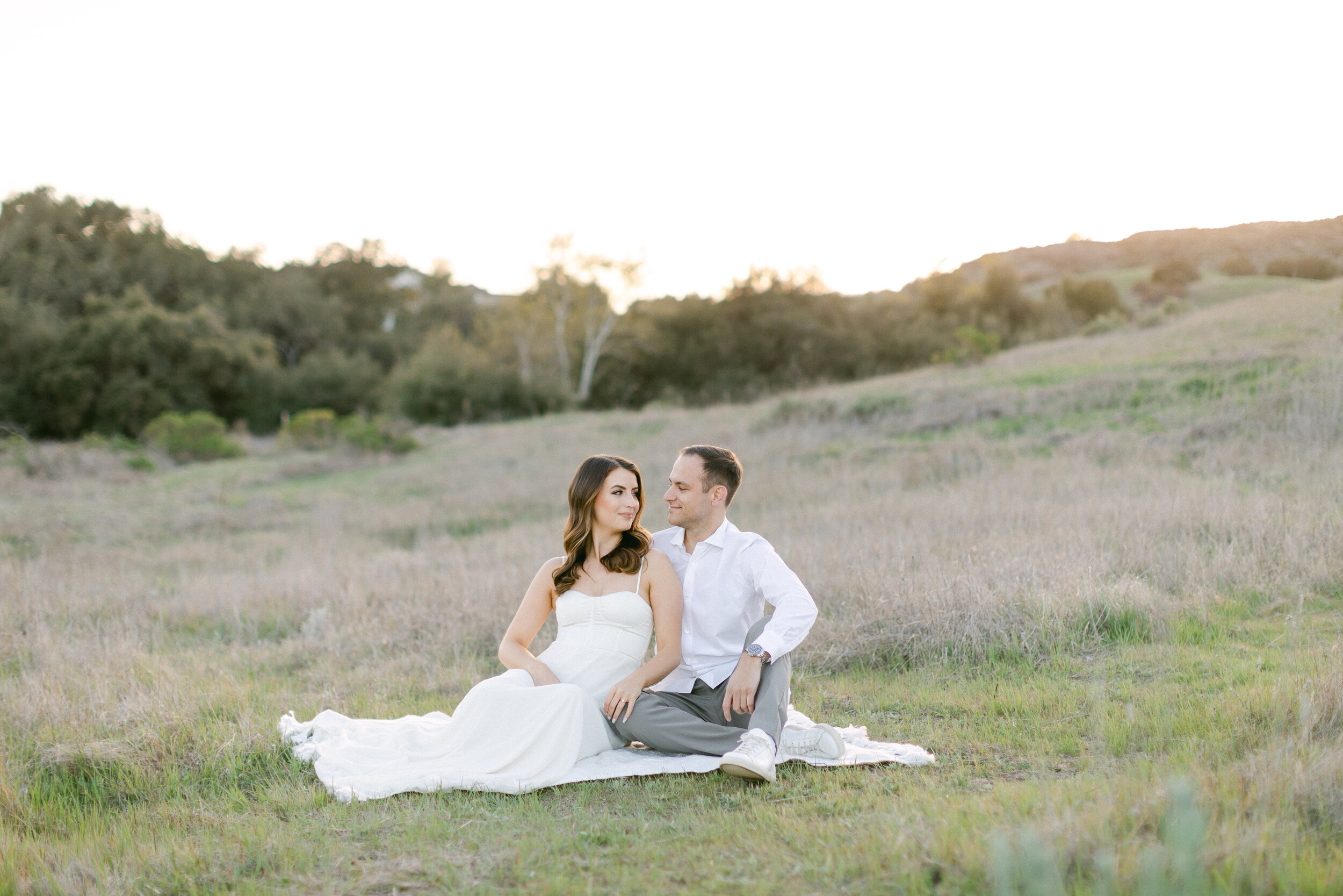 Brunette male and female sit on a blanket in a grassy field during their Orange County engagement photography session.