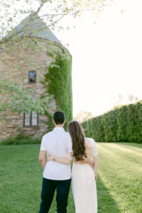Image shows bride and groom to be with their arms around each other's waist as they face away from the camera and look toward the beautiful, architectural Dovecote.