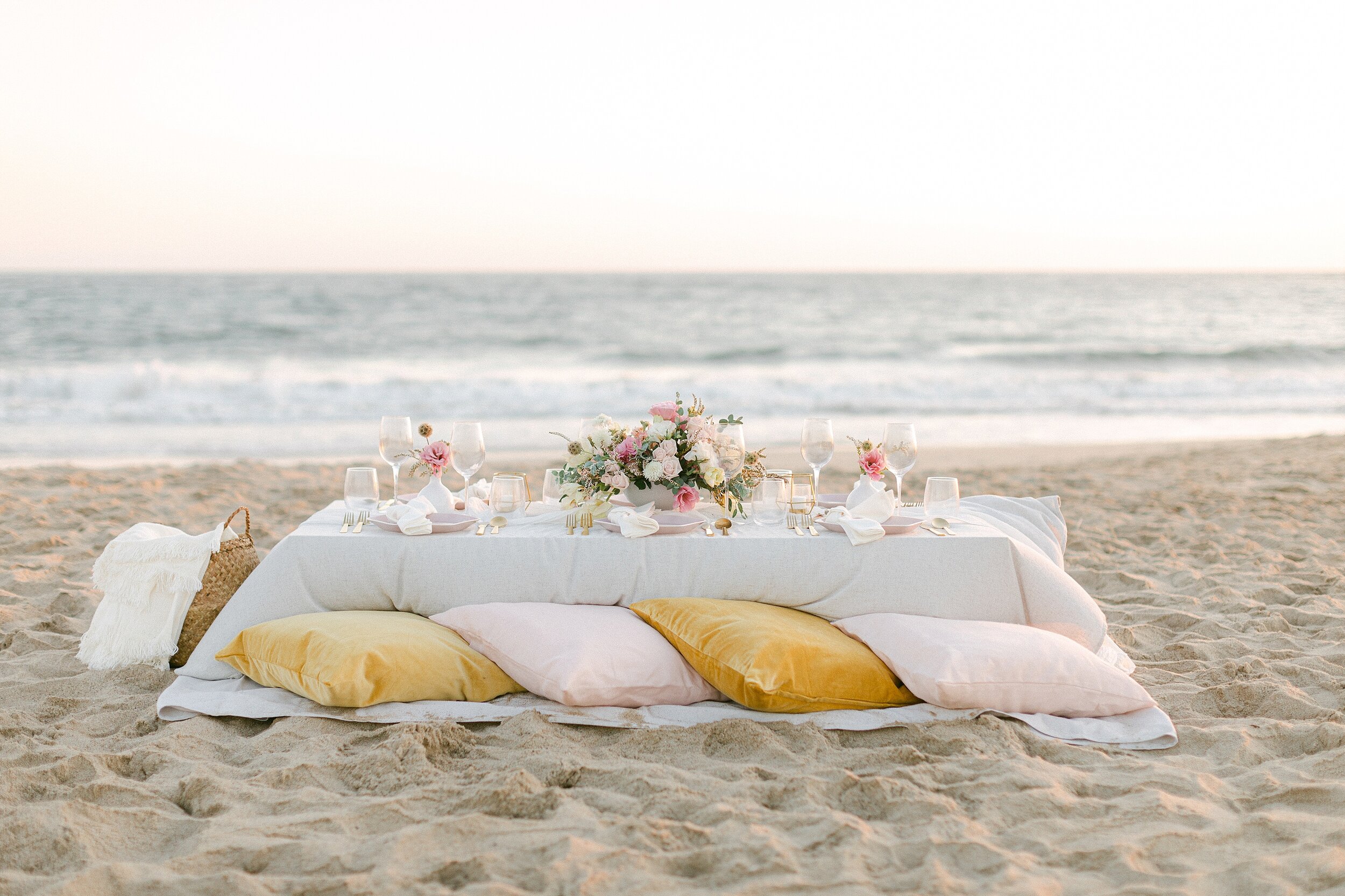 This beachside reception features a low table with a cream-colored linen table cloth, pink and white floral centerpiece, blush plates, gold flatware, and mustard yellow and blush pillow for seating.
