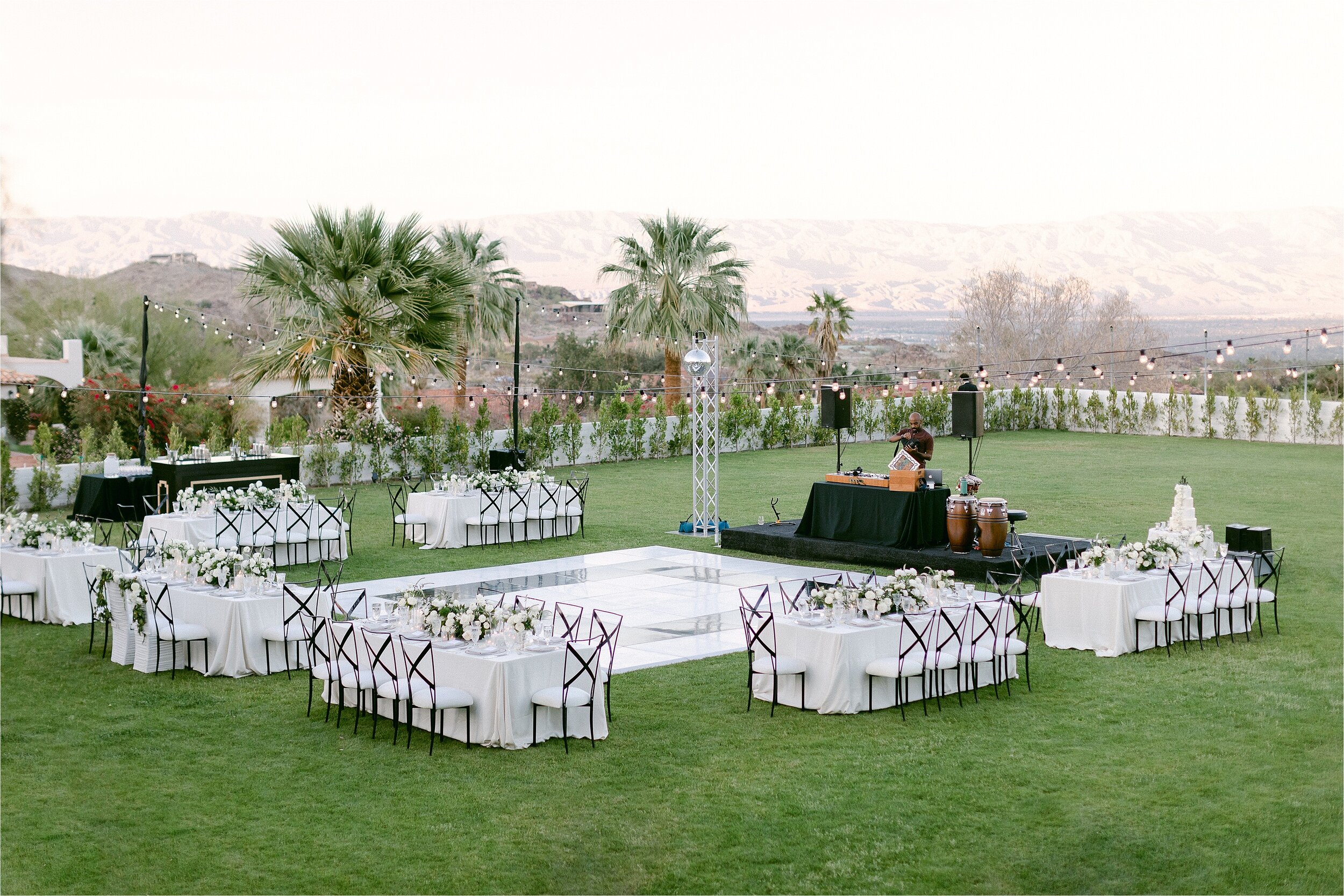 Elegant outdoor destination wedding reception at lush private estate in Palm Springs.  Featuring black and white dance floor, market lights, white linens, white floral centerpieces, and black metal chairs with white cushions. 