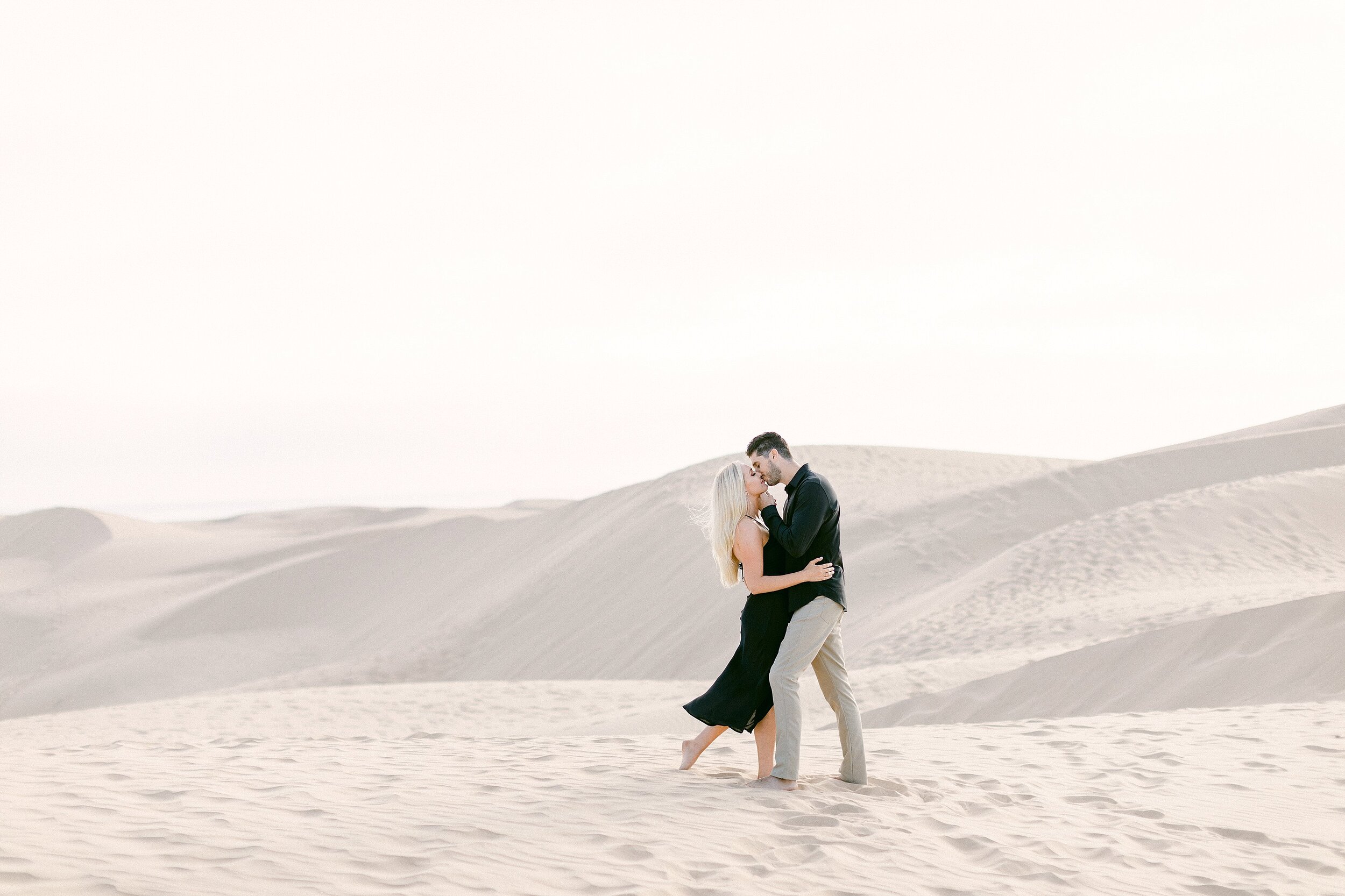 blonde female and brunette male share a kiss during their sand dunes engagement photos