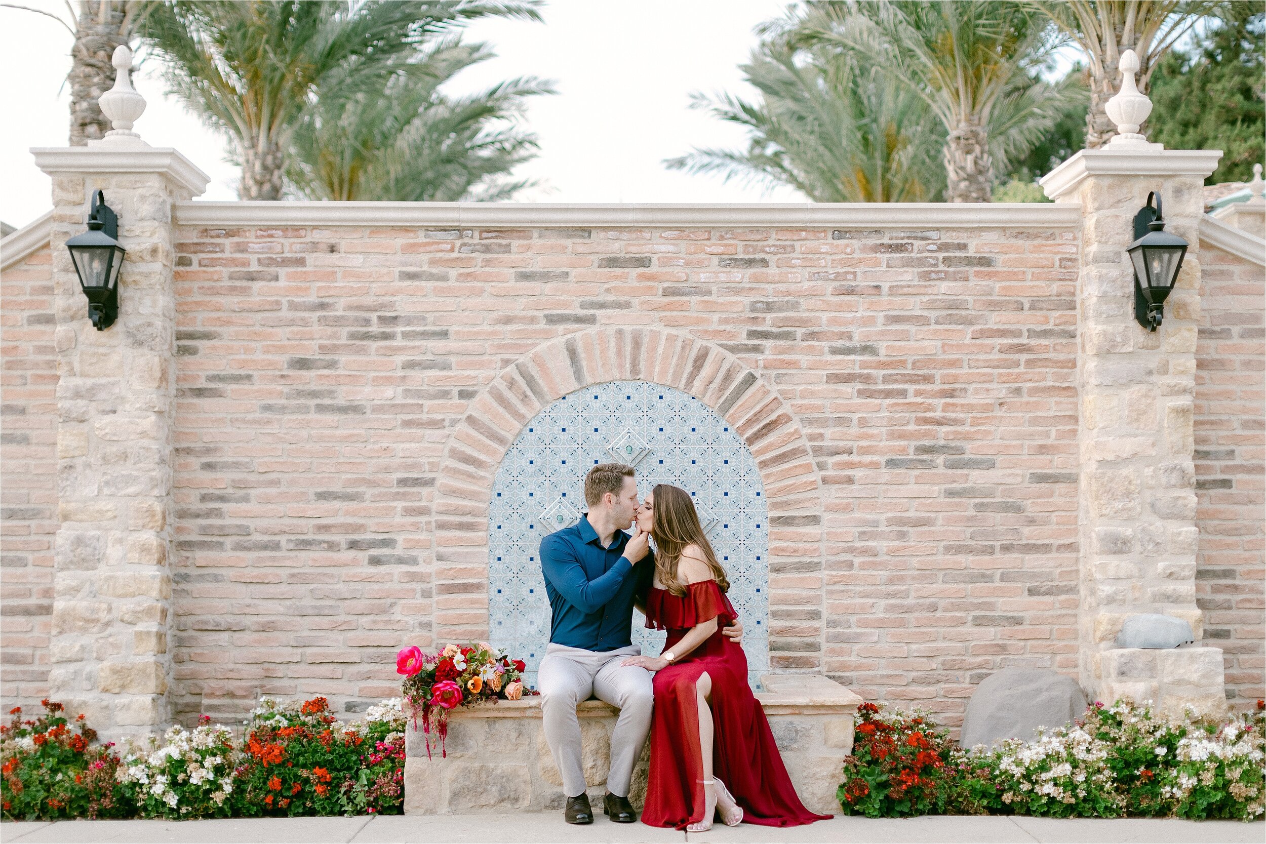 A couple sitting on the ledge of a fountain, kissing.  She is wearing a long, off shoulder wine colored dress with a leg slit.  He is wearing a midnight blue button down shirt with khaki pants.