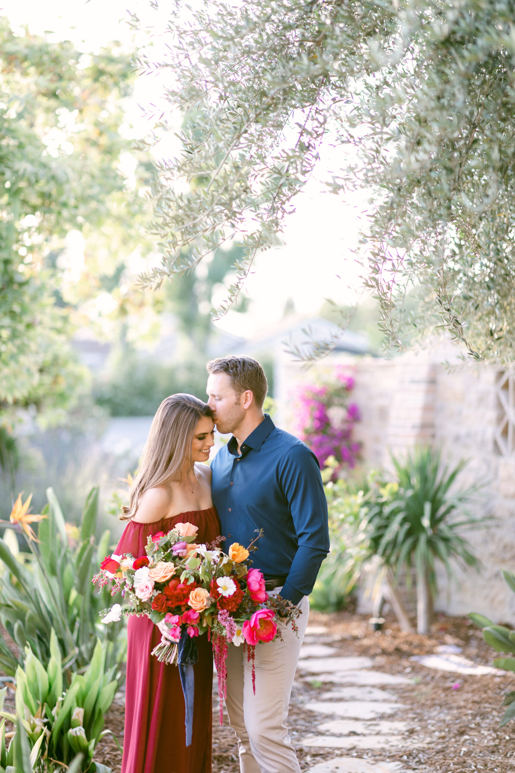 Colorful springtime engagement session featuring a brunette female wearing a long, off shoulder, wine colored maxi dress holds a large, vibrant bouquet as her fiance wearing a dark blue button down kisses her temple.