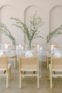 A wedding reception tablescape was created for a downtown Los Angeles editorial photo shoot. Image fetures a clear acrylic table with a cream-colored place-setting, topped with a cream-colored napkin and paper menu. Featuring olive branch centerpieces, cream-colored candles and whicker chairs.
