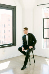 Full body portrait features the groom wearing a black tux, black bow tie and black shoes, sitting on a black metal stool while looking off into the distance.