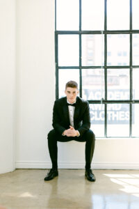 The groom sits on a window pane, wearing a black tux, black bow tie and black shoes while being photographed for an editorial wedding photography campaign in downtown Los Angeles.