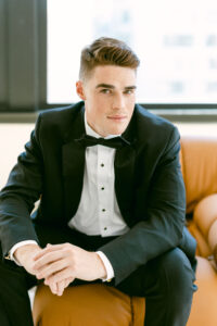 Groom poses for a portrait while sitting on a camel colored leather couch while wearing a black tux with a black bow tie.