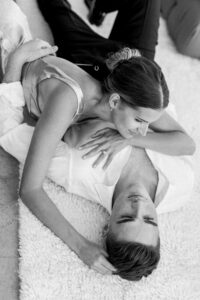Black and white photo showing a relaxed, intimate moment shared as the groom lays on a white rug and bride drapes her body over his while playing with his hair.