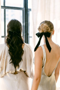2 bridesmaids facing away from the camera showcasing the backs of their champagne colored silk gowns and black velvet hair bows.