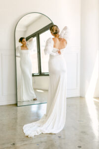 A blonde bride with her hair in a bun, looks in the mirror as she zips up her fitted, chiffon wedding gown.