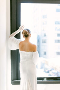 Blonde bride with her hair in a bun, rests her arm on a black window frame while looking out the window, thus showcasing the back of her delicate white wedding gown.