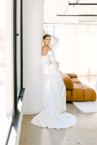 Full body photo shows the bride resting her back against a white pillar while placing one hand above her head. 