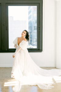 Bride sits on a stool in front of a large window wearing a delicate chiffon and lace bridal gown with her hair in a ponytail.