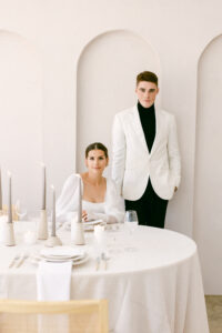 Bride sits at wedding reception table while the groom stands behind her. The bride is wearing a white bridal gown featuring puff sleeves and the groom is wearing black pants, a black turtleneck and a white suit jacket.
