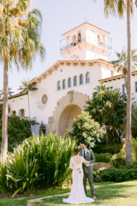 Scenic shot of brunette bride wearing a white lace long sleeved wedding dress has her back to the camera as she embraces her groom, facing toward the camera with black hair, wearing an olive green suit while standing on the lawn in front of the Santa Barbara Courthouse following their intimate micro wedding.