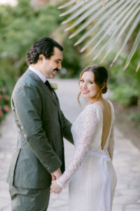 Brunette bride wearing a white lace long sleeved wedding dress, has her back to the camera as she looks over her shoulder while holding her groom's hand as he smiles at her. Groom has black hair, and is wearing an olive green suit while standing on a cobblestone pathway under large palm fronds at the Santa Barbara Courthouse.