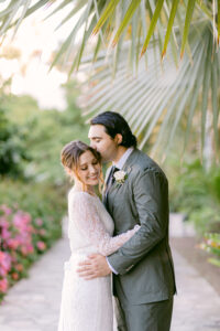 Brunette bride wearing a white lace long sleeved wedding dress, embraces her groom with black hair, wearing an olive green suit while standing on a cobblestone pathway under large palm fronds during their intimate micro wedding at the Santa Barbara Courthouse.