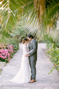 Brunette bride wearing a white lace long sleeved wedding dress, embraces her groom with black hair, wearing an olive green suit while resting their foreheads together while standing on a cobblestone pathway under large palm fronds during their intimate micro wedding at the Santa Barbara Courthouse.