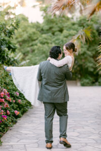 Brunette bride wearing a white lace long sleeved wedding dress, is swept off her feet and held by her groom with black hair, wearing an olive green suit while standing on a cobblestone pathway following their elopement at the Santa Barbara Courthouse.