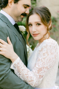 Close up image of a brunette bride wearing a white lace long sleeved wedding dress, looks straight at the camera while being embraced by her groom with black hair, wearing an olive green suit while he rests his head against his bride's temple following their micro wedding at the Santa Barbara Courthouse.