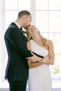Groom embraces his blonde bride and kisses her forehead as she snuggles into him and laughs.