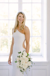 Portrait of blonde ride smiling toward her groom and wearing a white textured one-shouldered gown.