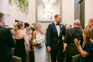 Photo of bride and groom smiling and greeting guests during their recessional immediately following their wedding ceremony as guests cheer and applaud.
