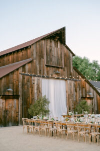 The iconic Greengate Ranch Barn provided the perfect backdrop for Brian and Shannon's outdoor wedding reception with long tables, light wood, crossback chairs, beautiful florals and hanging market lights.