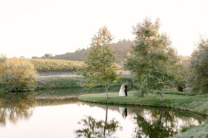 Horizontal image showcasing the beautiful lake at Greengate Ranch surrounded by trees and vineyards as bride and groom embrace and their reflection is seen in the lake.