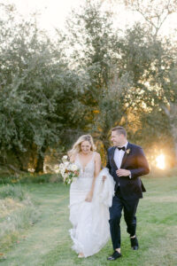 Bride and groom excitedly run toward their wedding reception as he holds her train while the sun sets behind them.