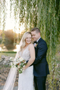 Brunette groom in a navy suit, embraces his bride as she nestles into him under a whispy willow tree.