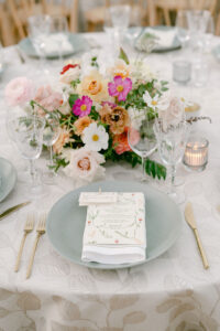 Colorful tablescape with beautifully textured linens, light blue plates, custom farm to table menus, vibrant florals, and etched glassware.