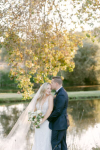 Groom kisses bride's forehead under a tree canopy with the lake in the background, as her veil cascades over her shoulder and she holds her bouquet at her waist.