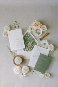 Wedding invitation flatlay showcasing the sage green invitation accents complimented by florals, vintage stamps and the couple's wedding rings.