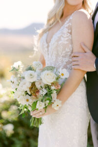 Detail photo of white and light pink bridal bouquet as bride leans against her groom and holds her bouquet at her waist.