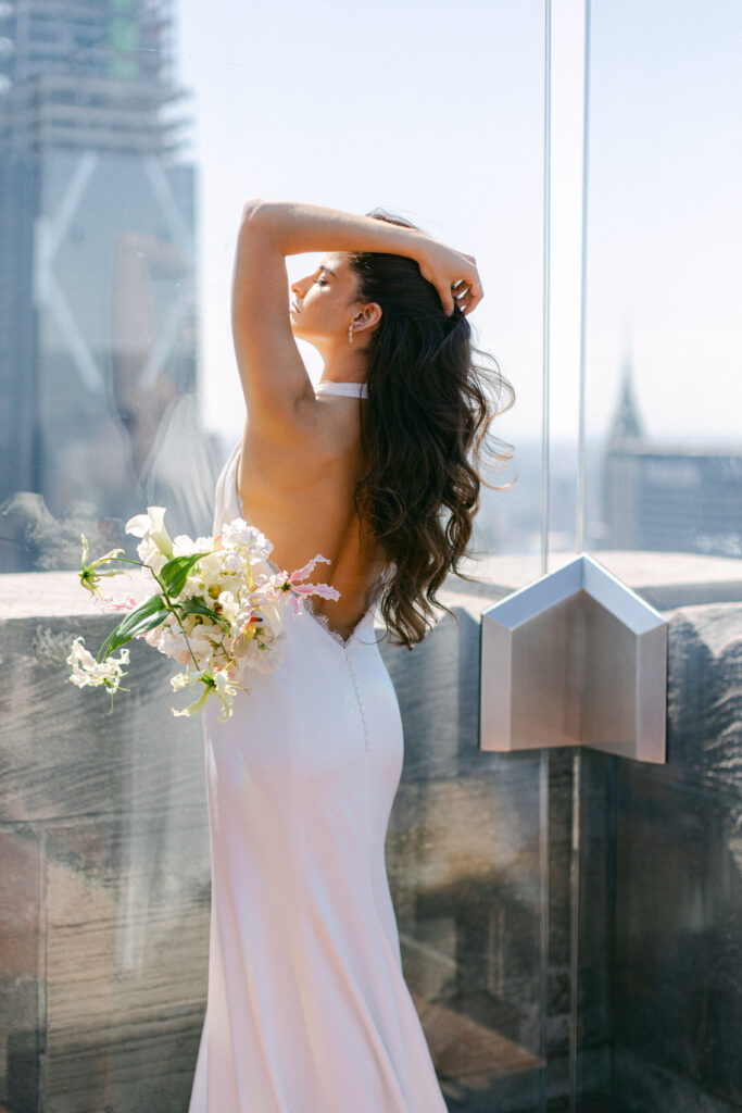 Bride delicately runs her hand through her hair, showcasing her backless, halter sheath gown.